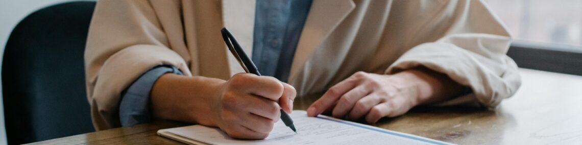 A woman in a formal setting fills out paperwork on a clipboard at an office desk.